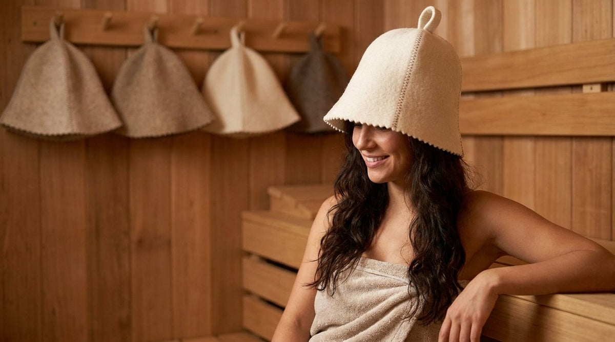 Woman with long healthy hair sitting in a wooden sauna wearing a white wool sauna hat, relaxed, warm tones